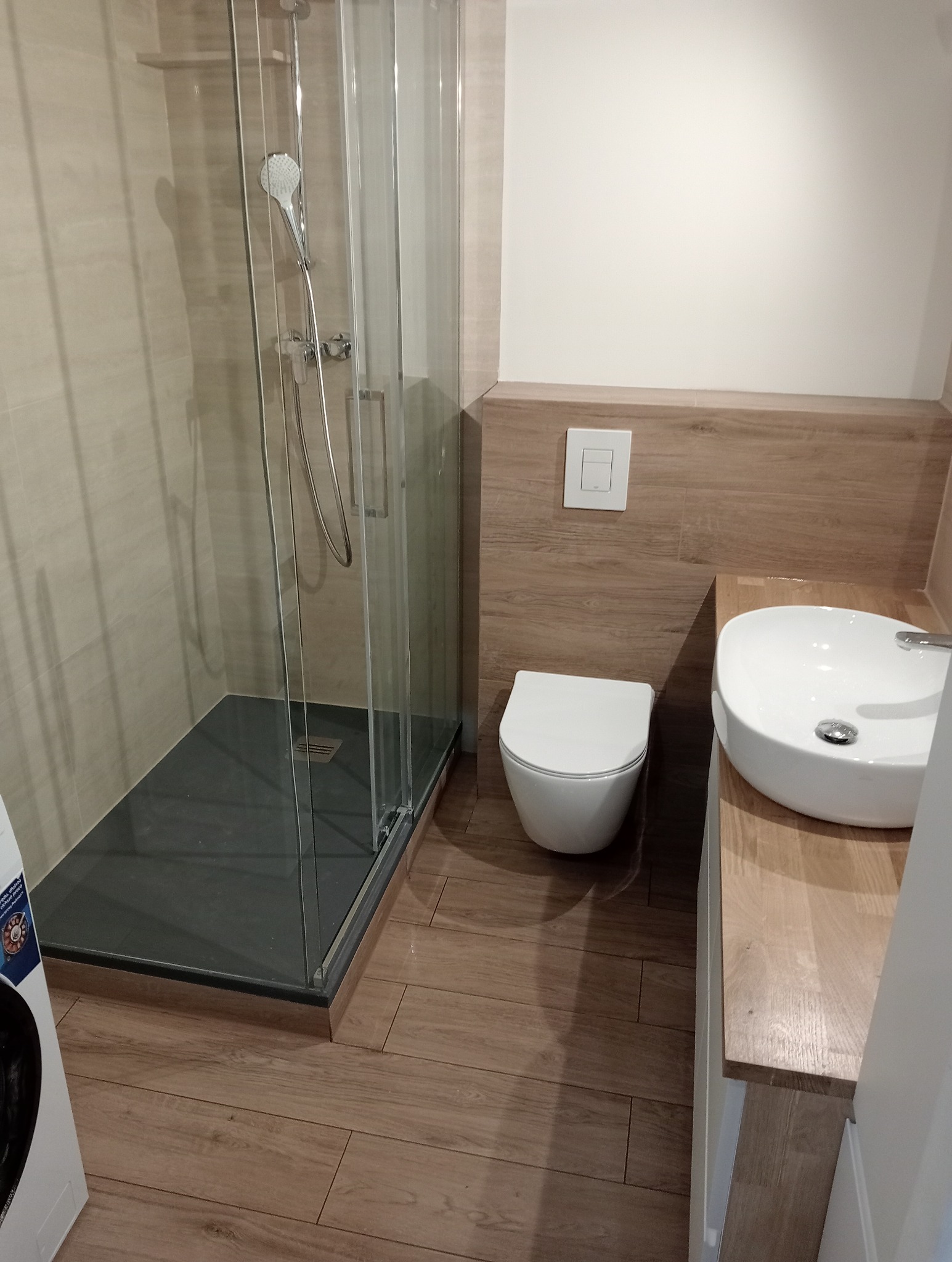 Nowoczesna łazienka z kabiną prysznicową typu walk-in, podwieszaną toaletą i umywalką nablatową. Płytki imitujące drewno na podłodze i części ścian.