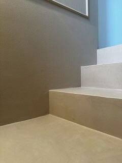 Minimalistyczne schody z mikrocementu w odcieniach beżu i szarości, z akcentem błękitu na ścianie. Gładka powierzchnia, nowoczesny design, widok z dołu, oświetlenie naturalne.