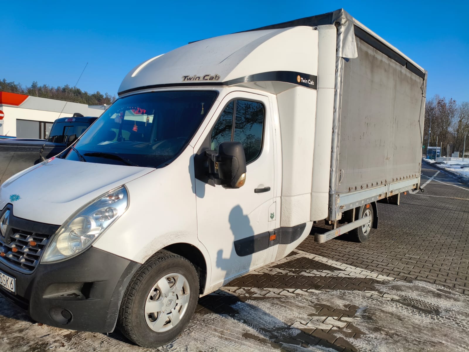 Biały bus Renault Master Twin Cab z plandeką na parkingu w słoneczny dzień. Widoczne ślady śniegu na kostce brukowej. Transport busem.