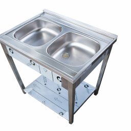 Gastro-Meble Agnieszka Moś-Wyrobek - Stalowy zlew dwukomorowy na metalowym stelażu z półką, widok z góry pod kątem. Zlew o prostych kształtach, z widocznymi elementami montażowymi i folią ochronną.