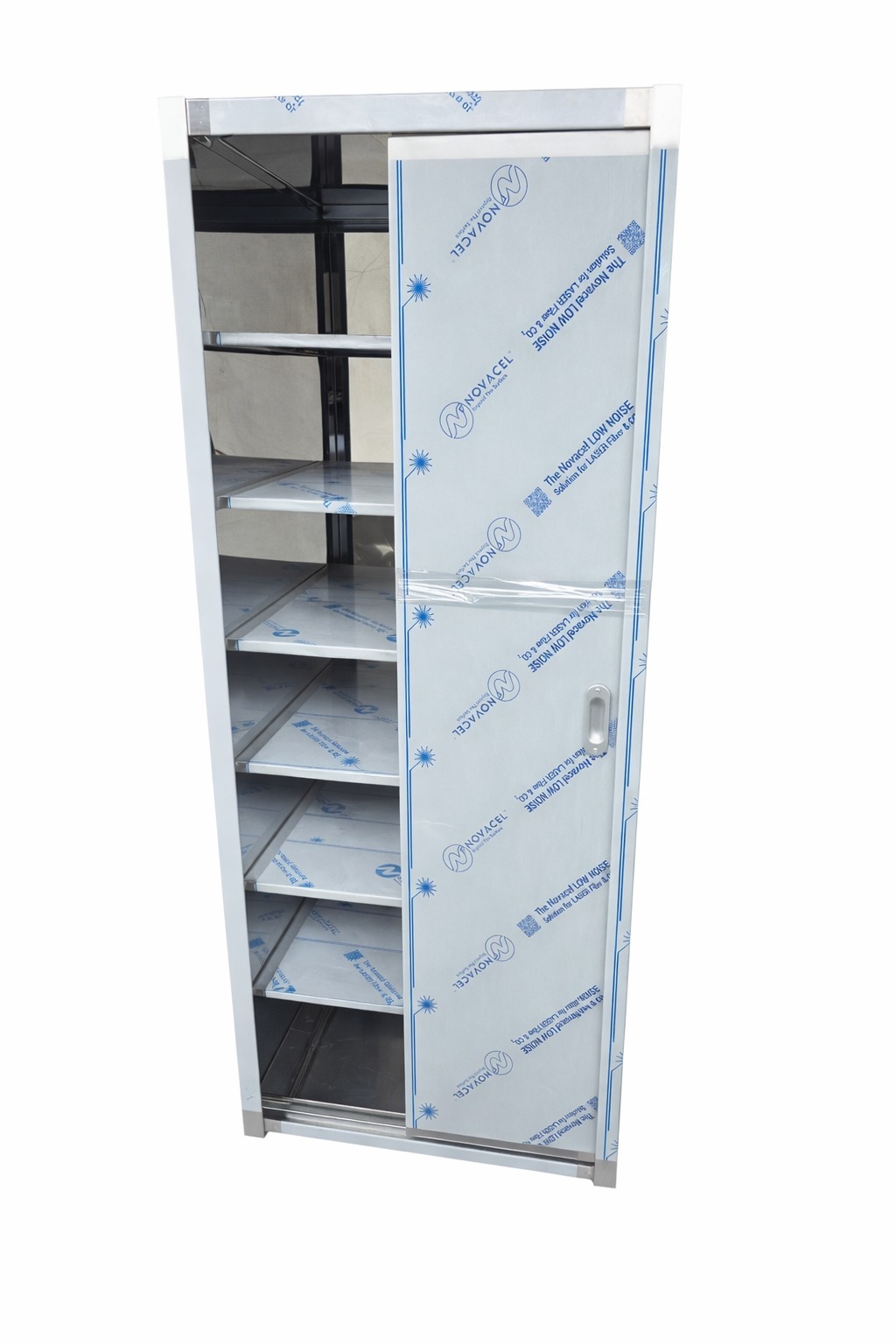 Metalowa szafka kuchenna z przesuwnymi drzwiami, jeszcze w folii ochronnej, z widocznymi półkami wewnątrz. Nowoczesny design, surowy materiał.