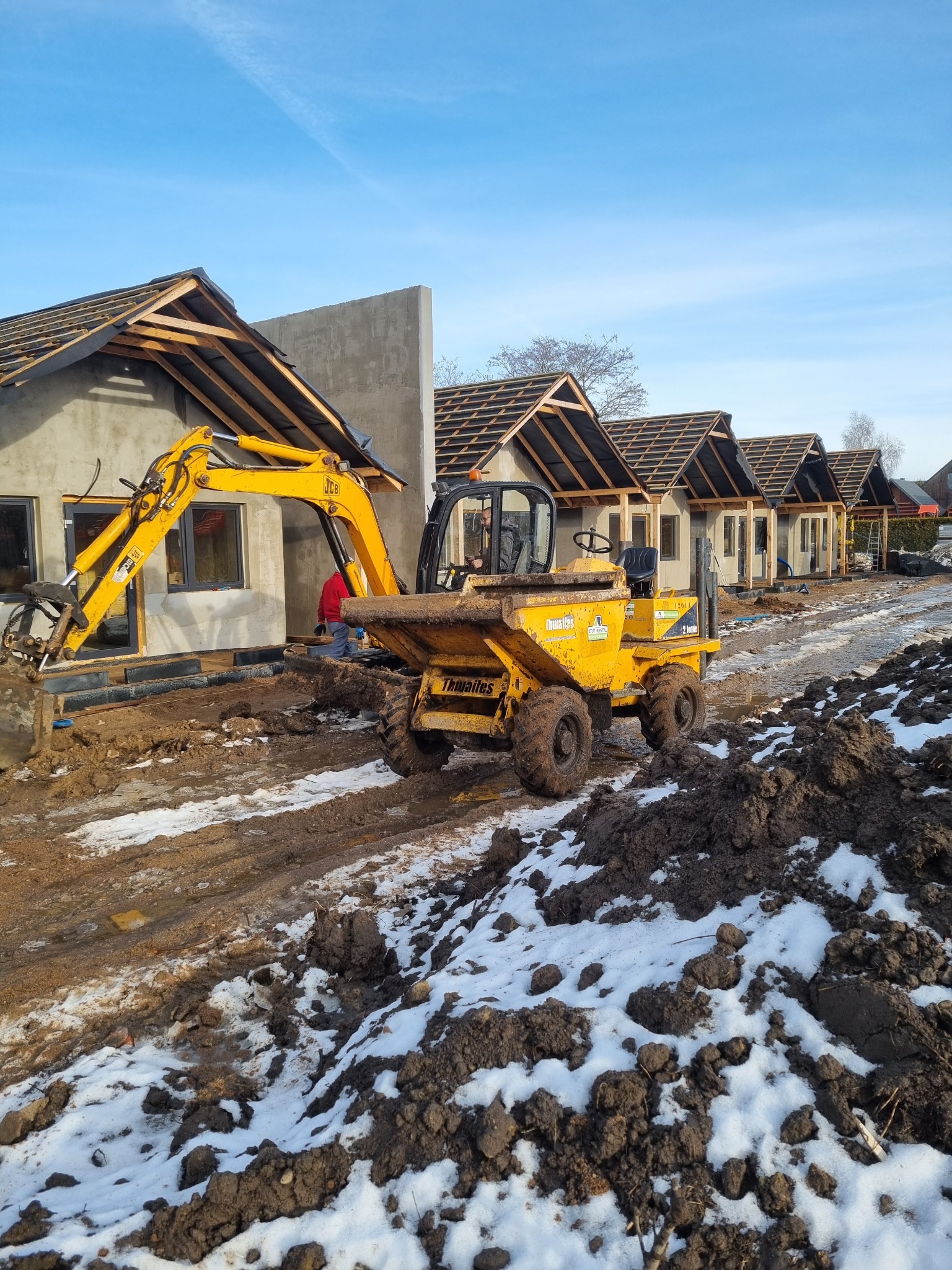 Żółta koparka JCB i wywrotka Thwaites na placu budowy szeregu domów w stanie surowym otwartym, z ziemią i resztkami śniegu. Błękitne niebo w tle.