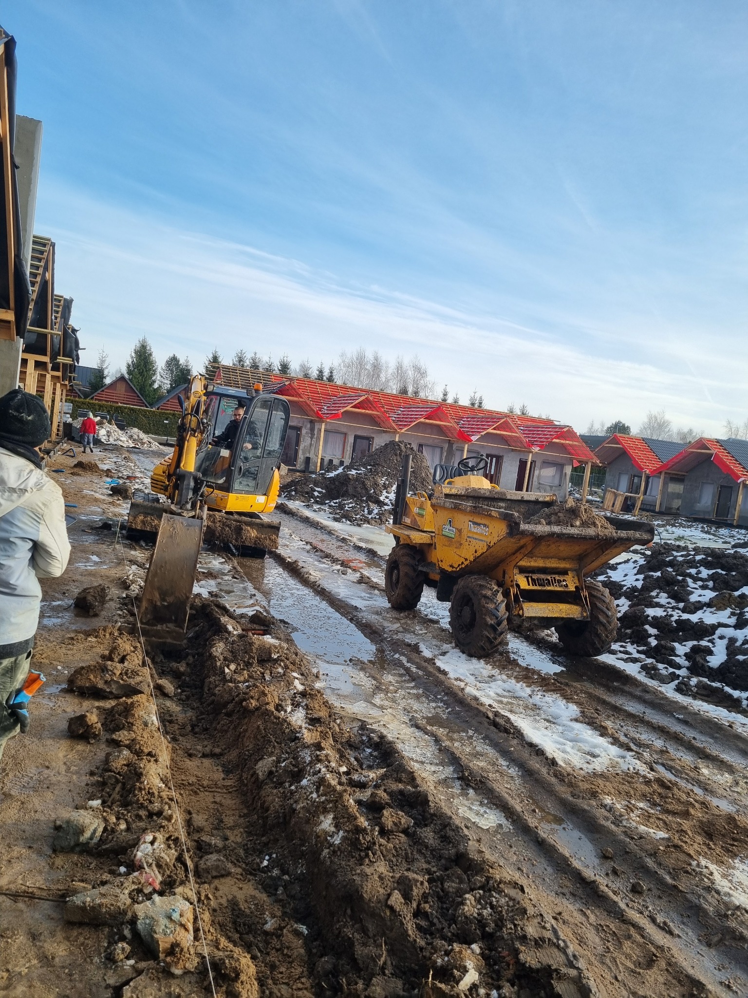 Żółta koparka i wywrotka na błotnistym placu budowy z widokiem na domy w trakcie budowy z czerwonymi dachami i błękitne niebo w tle.