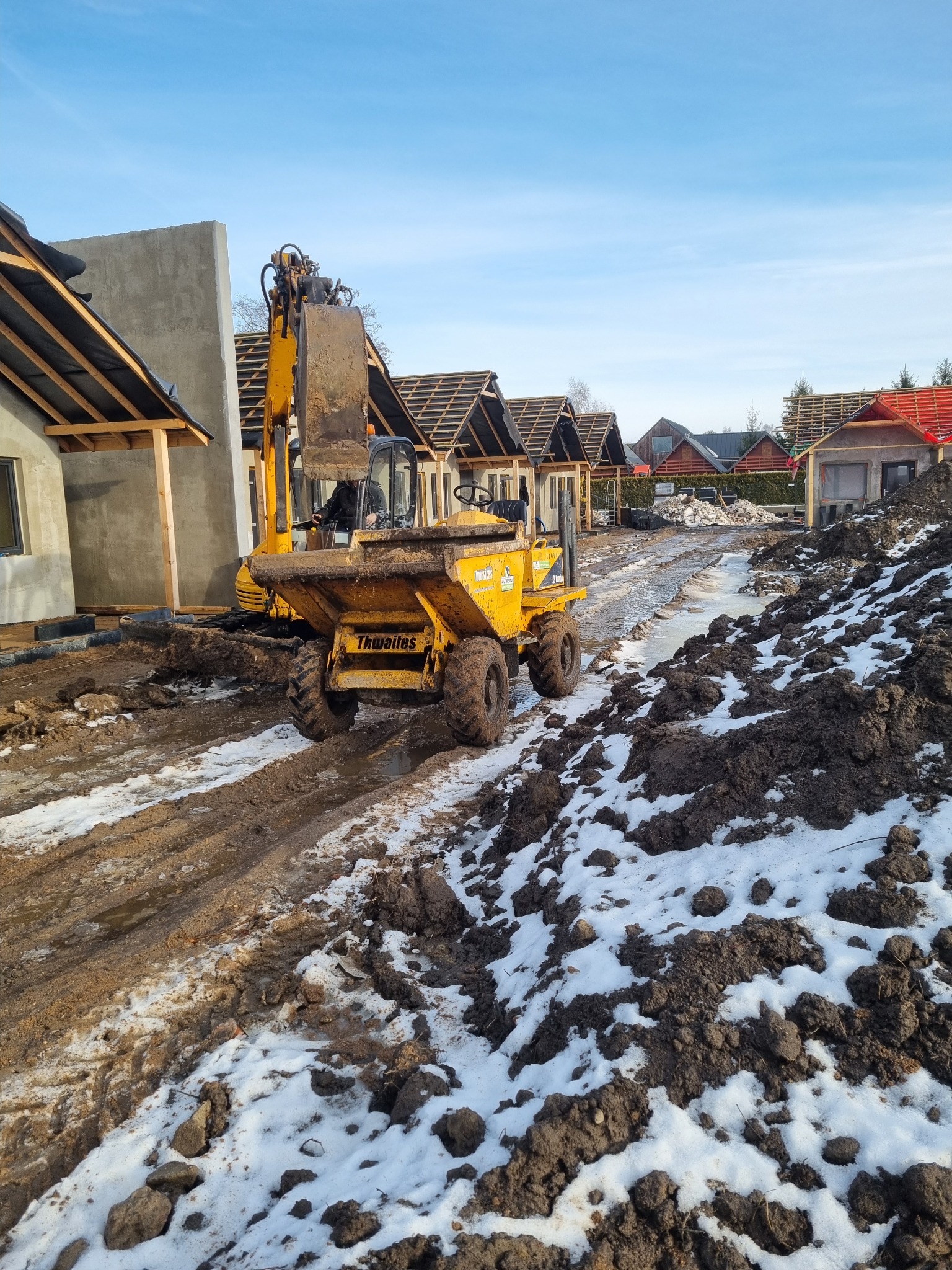 Żółta koparka Thwaites na placu budowy z ziemią i śniegiem, w tle budynki w stanie surowym otwartym, błękitne niebo. Prace ziemne w toku.