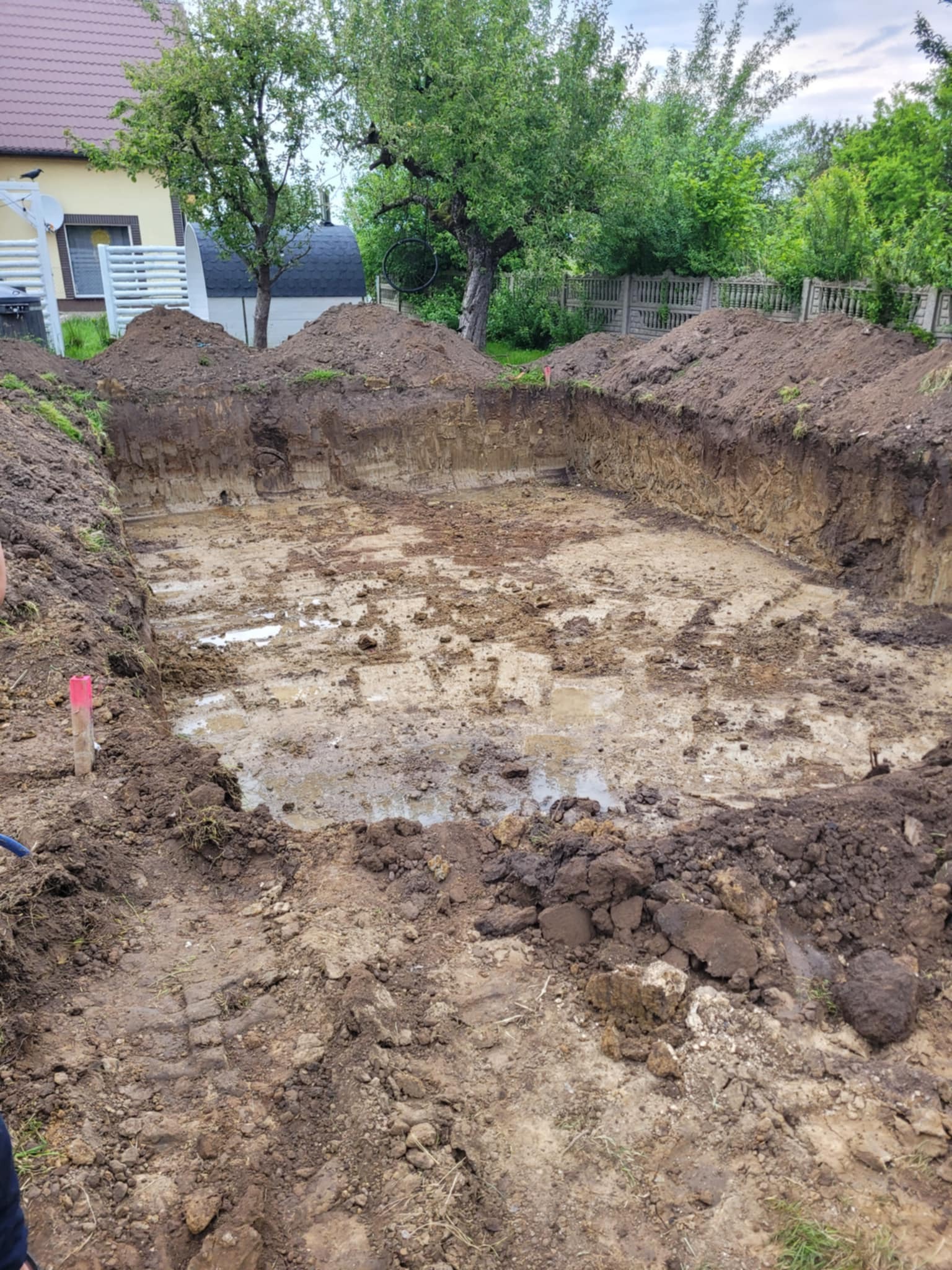 Prostokątny wykop w ziemi, prawdopodobnie pod fundamenty, z widocznymi śladami wody i błota na dnie. W tle dom i drzewa, co sugeruje teren budowlany.