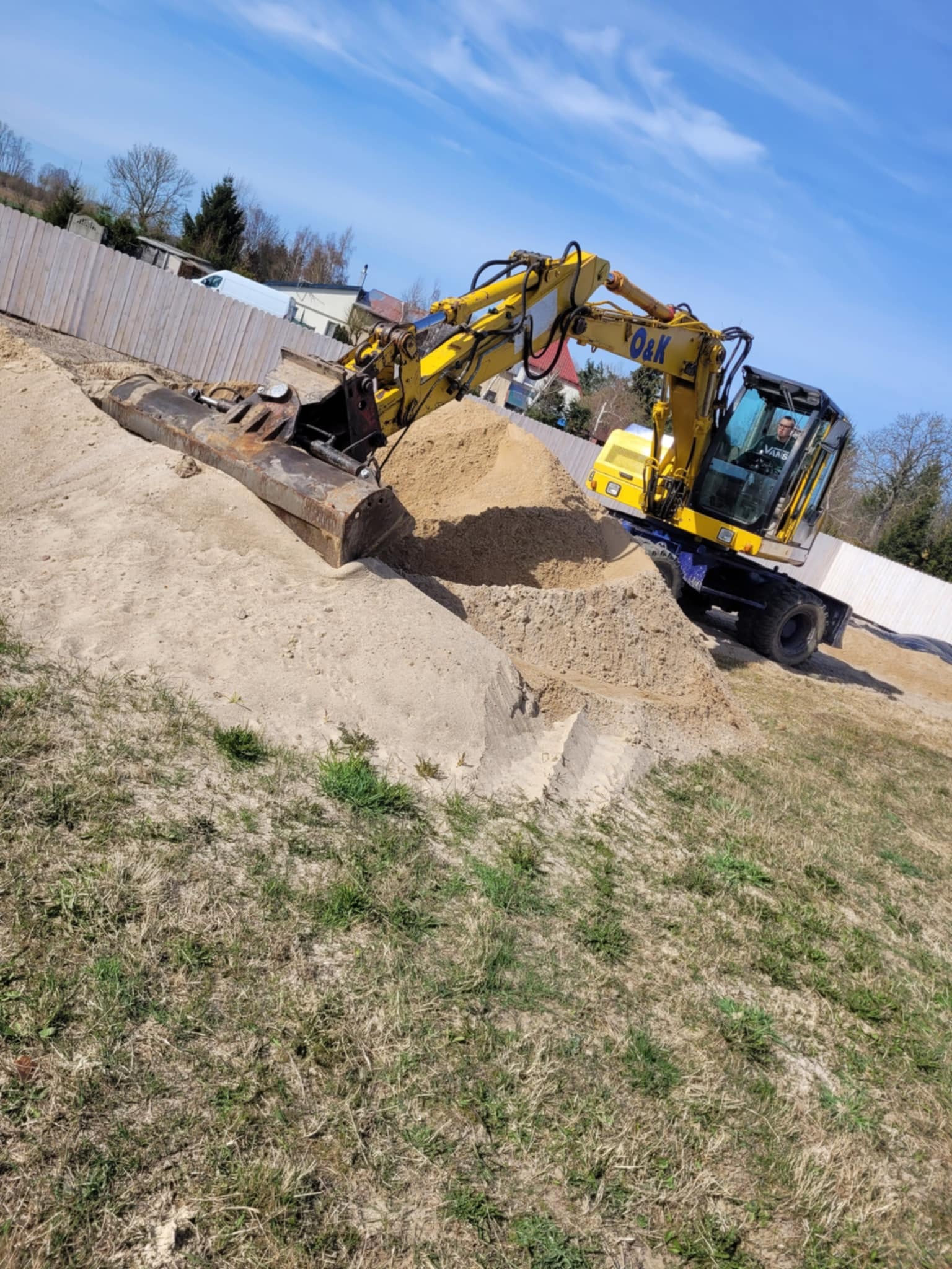 Żółto-czarny kołowy koparka O&K nabiera piasek na tle błękitnego nieba i drewnianego ogrodzenia. Operator w kabinie. Widoczny teren z trawą.
