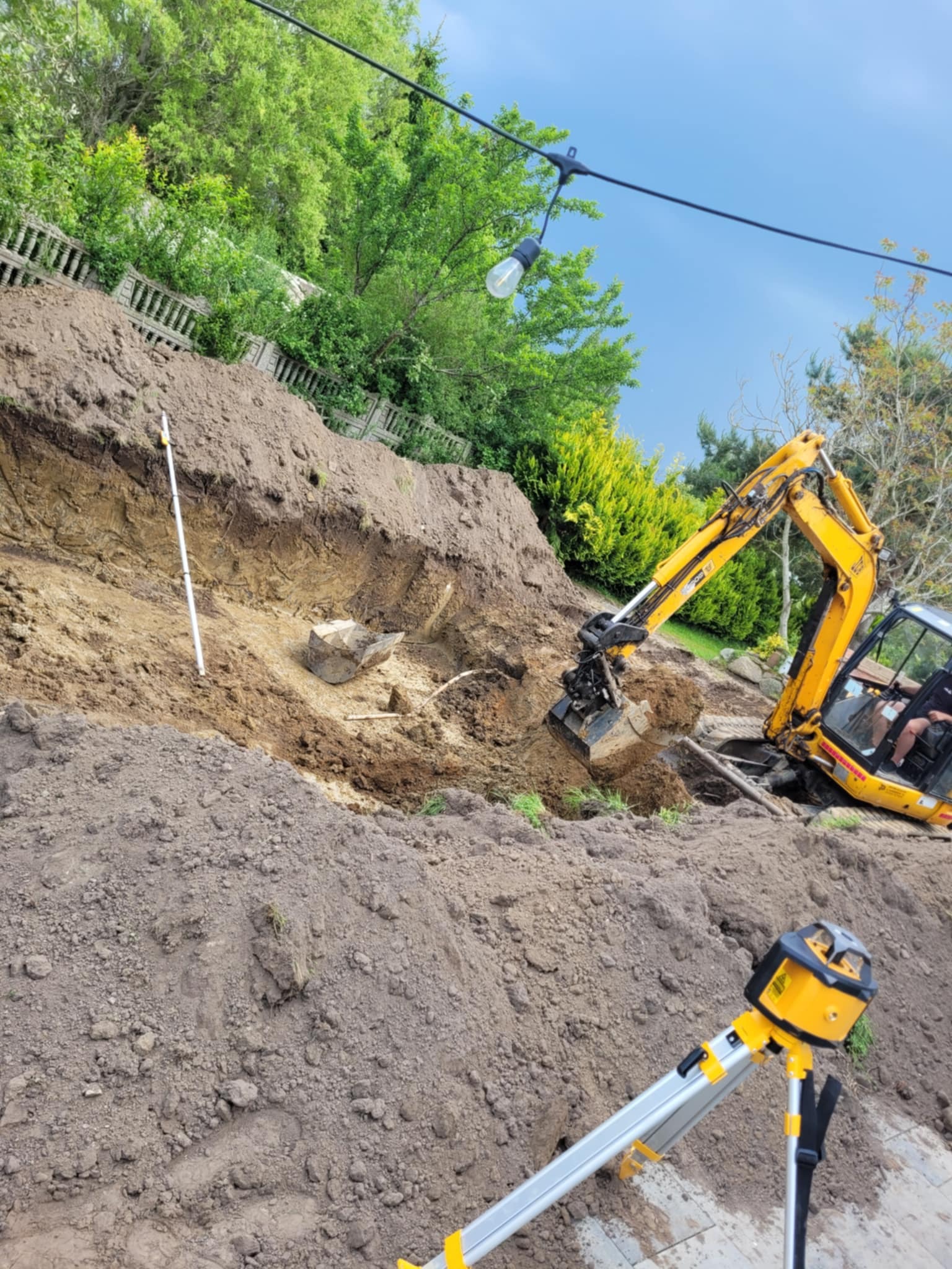 Żółta koparka JCB w trakcie wykopu ziemi na zboczu, widoczny niwelator laserowy i łata niwelacyjna. Prace ziemne w ogrodzie.