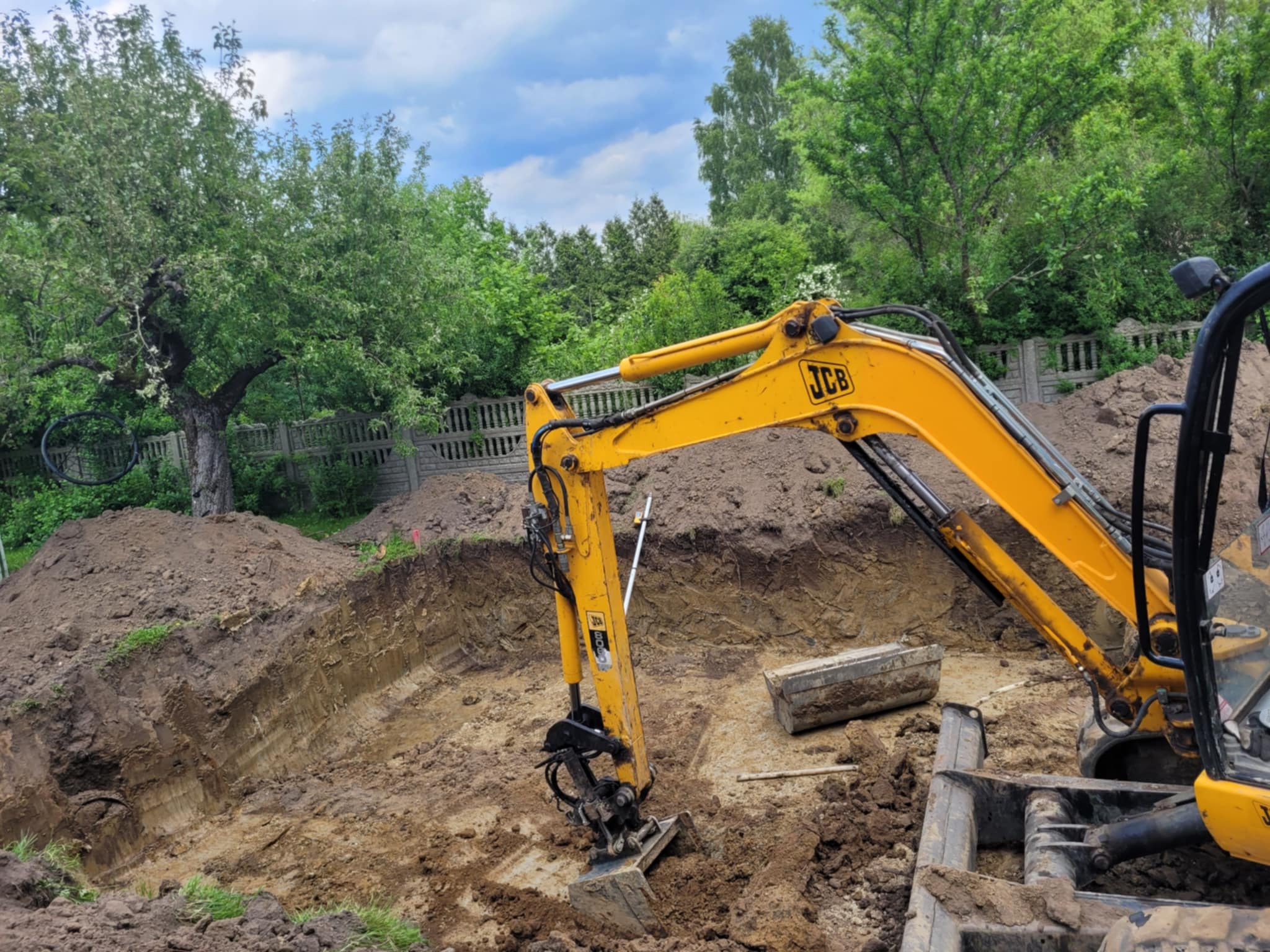 Żółta koparka JCB w wykopie ziemnym na tle zieleni i błękitnego nieba. Widoczny fragment ogrodzenia i drzewo. Prace ziemne w toku.