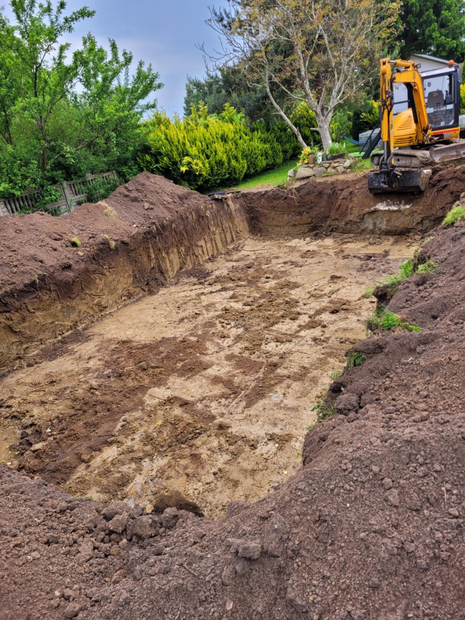 Żółta koparka przy wykopanym stawie w ogrodzie. Widoczne ślady opon na dnie wykopu i pryzma ziemi na brzegu. Trawa i krzewy w tle.