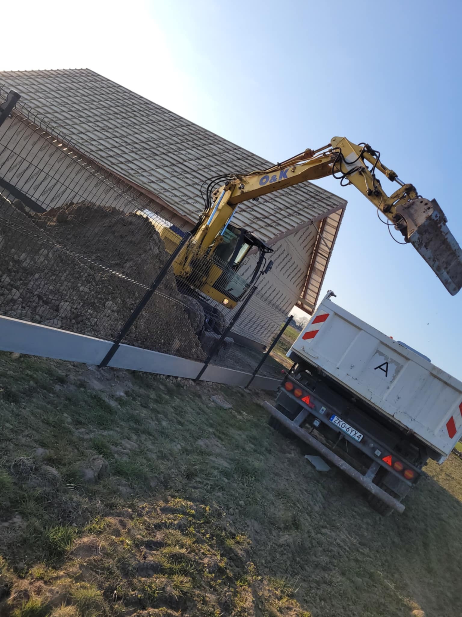 Żółta koparka O&K załaduje ziemię na białą wywrotkę na tle ogrodzenia i budynku w trakcie budowy. Ujęcie z dołu, obrócone w prawo.