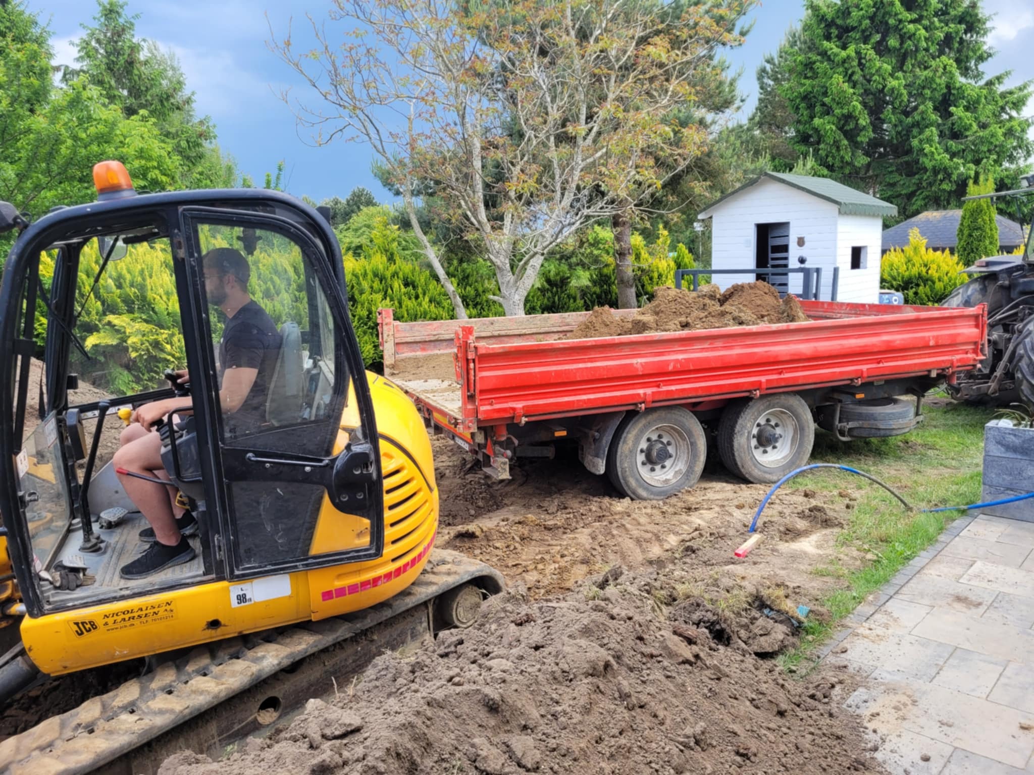 Żółta mini koparka JCB z operatorem w kabinie, ciągnąca czerwoną przyczepę z ziemią na tle zieleni i białego domku. Wykopany rów w ziemi.