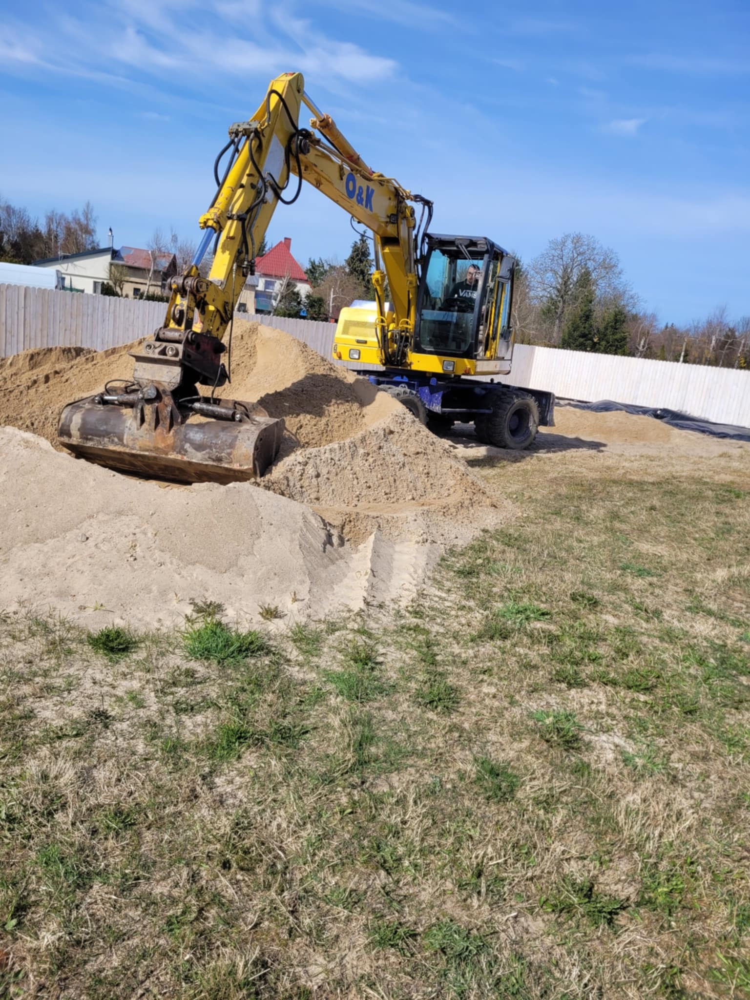Żółta koparka O&K na placu budowy, z wykopanym dołem i stertą piasku. W tle białe ogrodzenie i budynki pod błękitnym niebem.