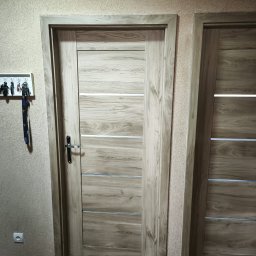 4Trade Paweł Milik - Dwa drewniane drzwi wewnętrzne z poziomymi, srebrnymi wstawkami. Na ścianie wieszak na klucze. Widoczny fragment gniazdka elektrycznego i podłogi.
