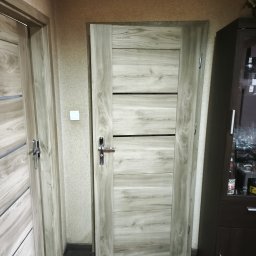 4Trade Paweł Milik - Zamknięte, drewniane drzwi wewnętrzne z poziomymi, czarnymi wstawkami, widoczne w korytarzu. Obok fragment szafki i drugich drzwi. Neutralna kolorystyka.