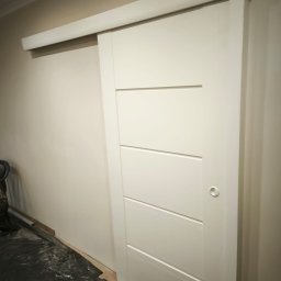 4Trade Paweł Milik - Białe, przesuwne drzwi z poziomymi frezami, zamontowane w jasnym wnętrzu. Widoczna listwa prowadząca i fragment podłogi zabezpieczonej folią malarską.