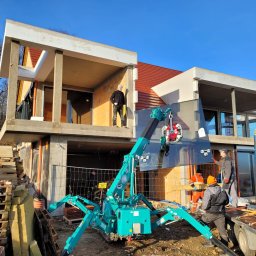 4Trade Paweł Milik - Montaż dużej szyby okiennej za pomocą specjalistycznego dźwigu na budowie domu. Widoczni pracownicy w strojach roboczych. Słoneczny dzień.