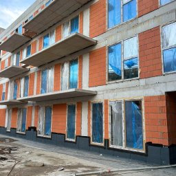 4Trade Paweł Milik - Budynek w trakcie montażu okien, zabezpieczonych folią. Widoczne elementy konstrukcyjne i materiały budowlane. Ujęcie z dołu, w tle błękitne niebo.