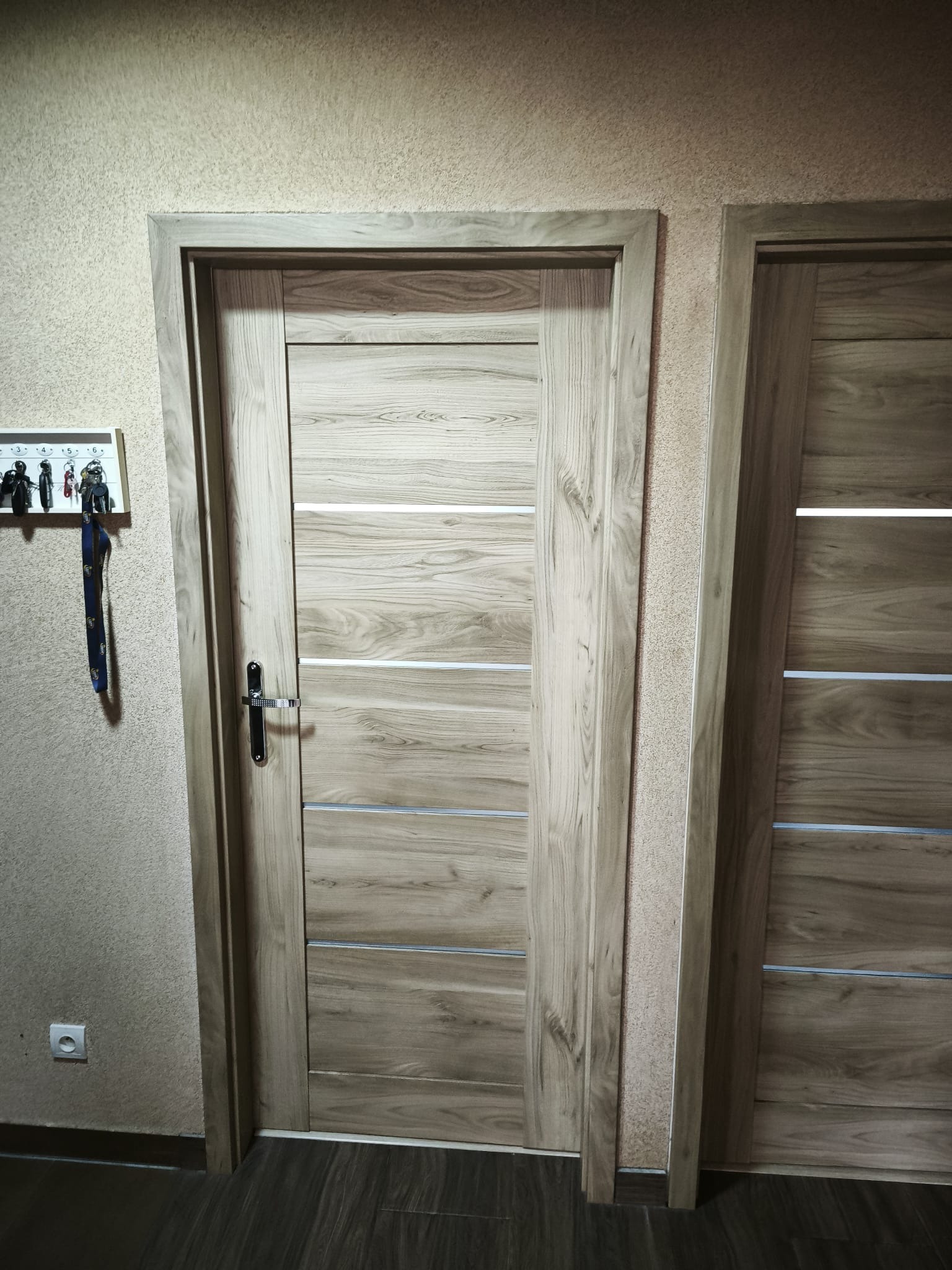 Dwa drewniane drzwi wewnętrzne z poziomymi, srebrnymi wstawkami. Na ścianie wieszak na klucze. Widoczny fragment gniazdka elektrycznego i podłogi.