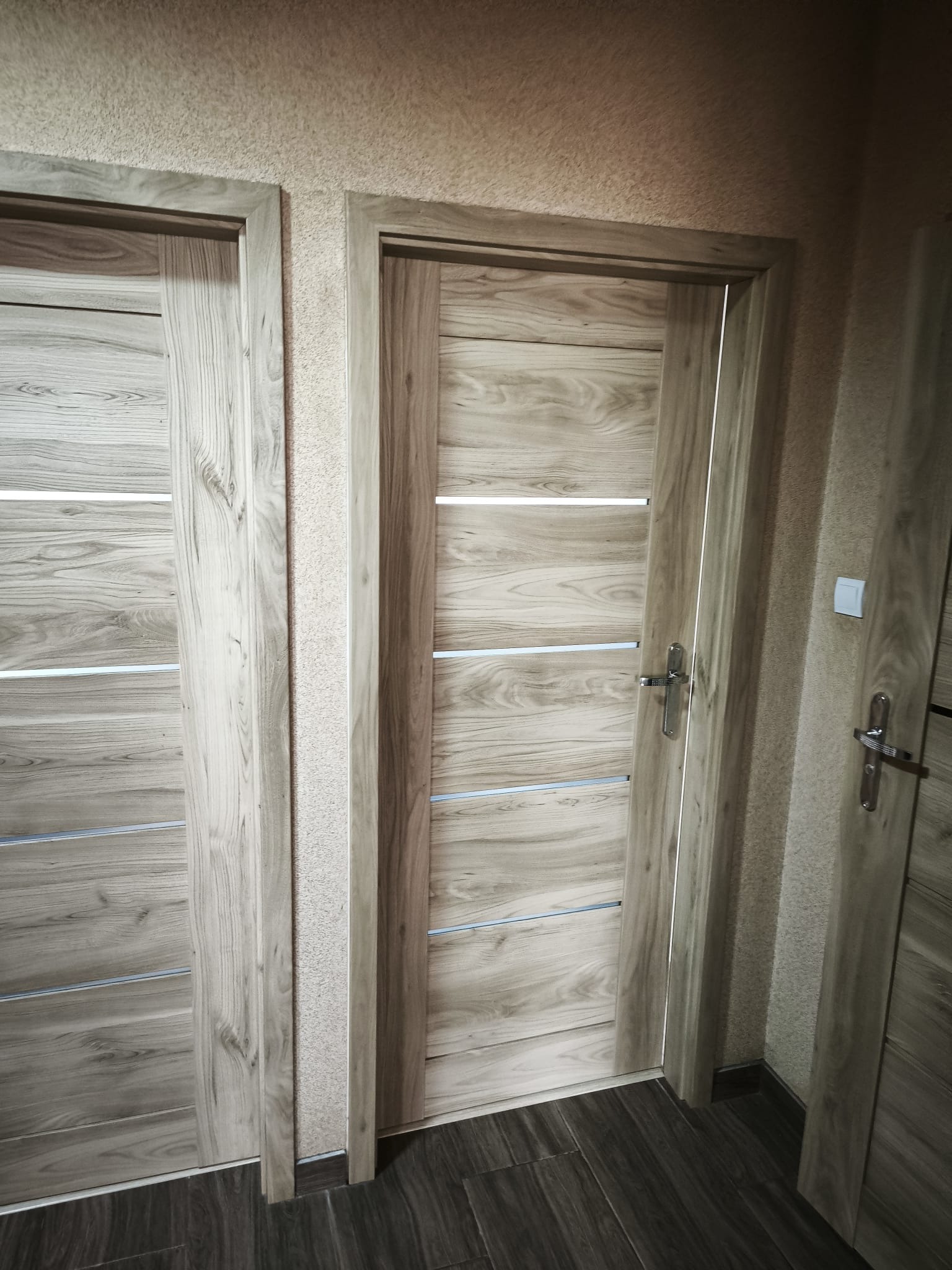Zamknięte, drewnopodobne drzwi wewnętrzne z poziomymi, srebrnymi wstawkami, widoczne w kadrze obok innych drzwi o podobnym designie, na tle teksturowanej ściany.