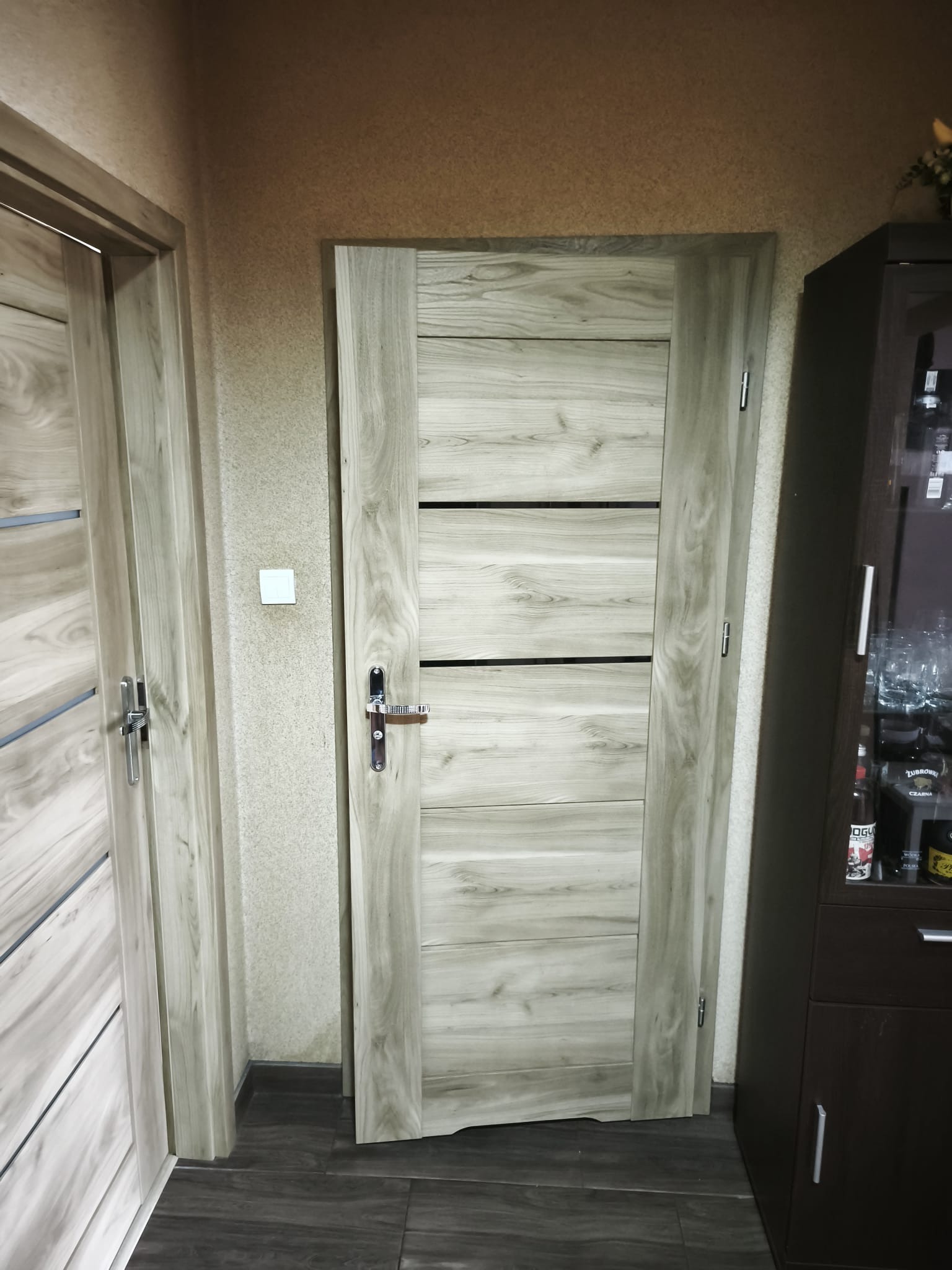 Zamknięte, drewniane drzwi wewnętrzne z poziomymi, czarnymi wstawkami, widoczne w korytarzu. Obok fragment szafki i drugich drzwi. Neutralna kolorystyka.