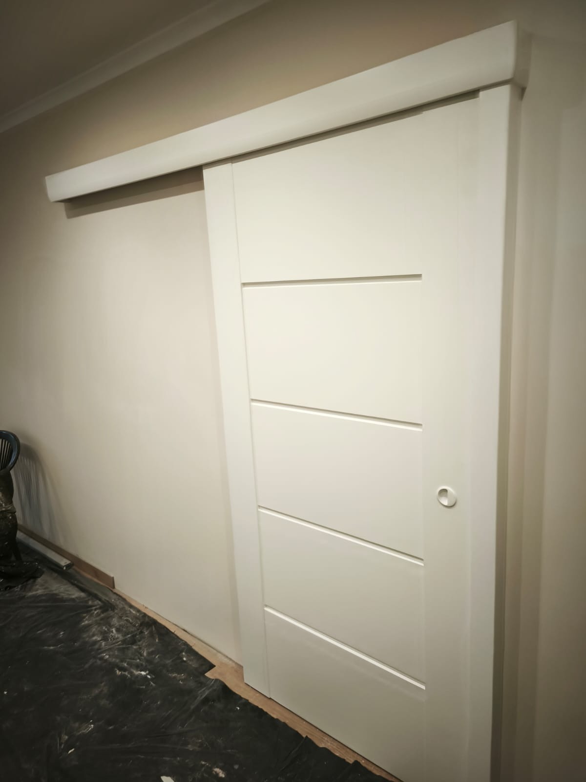 Białe, przesuwne drzwi z poziomymi frezami, zamontowane w jasnym wnętrzu. Widoczna listwa prowadząca i fragment podłogi zabezpieczonej folią malarską.