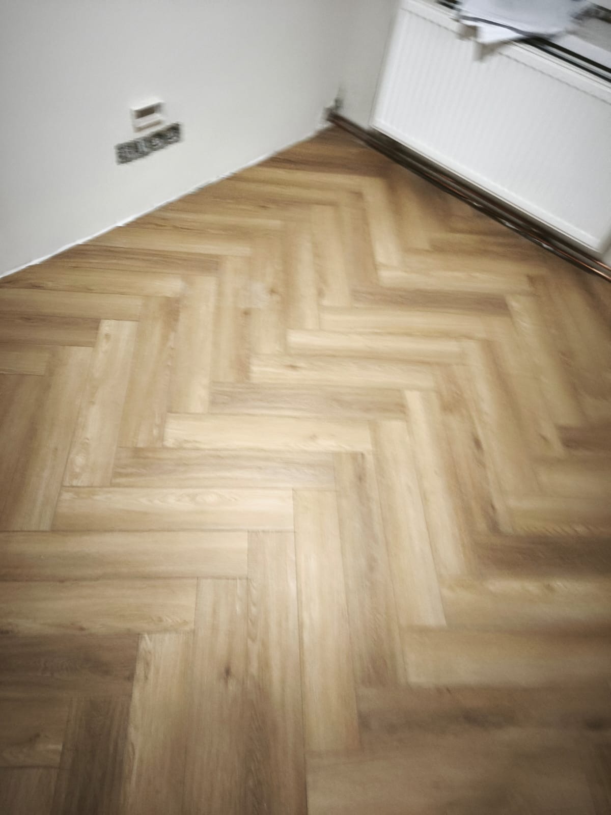 Ułożona podłoga z paneli w jodełkę, widok z góry, fragment ściany z gniazdkiem i grzejnik. Jasne panele, ciepłe oświetlenie.