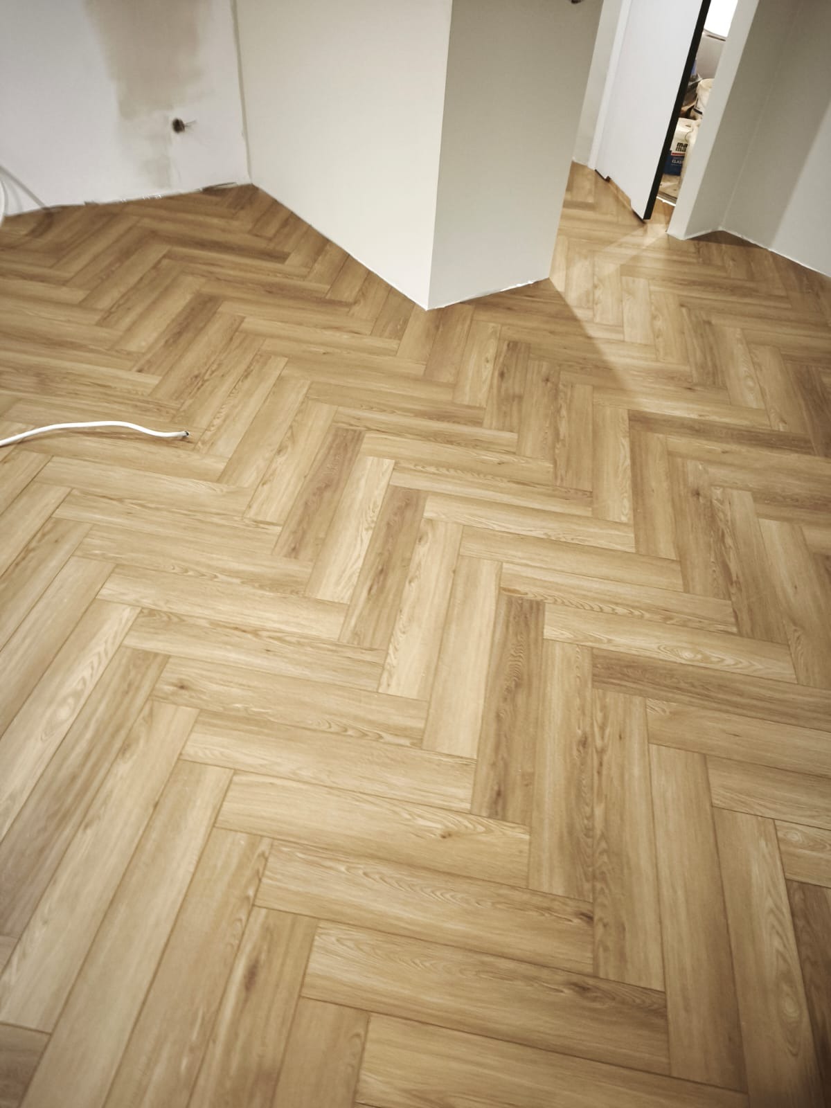 Ułożona podłoga z paneli w jodełkę, jasne drewno, widok z góry. Białe ściany i fragment otwartych drzwi w tle. Kabel elektryczny wzdłuż ściany.