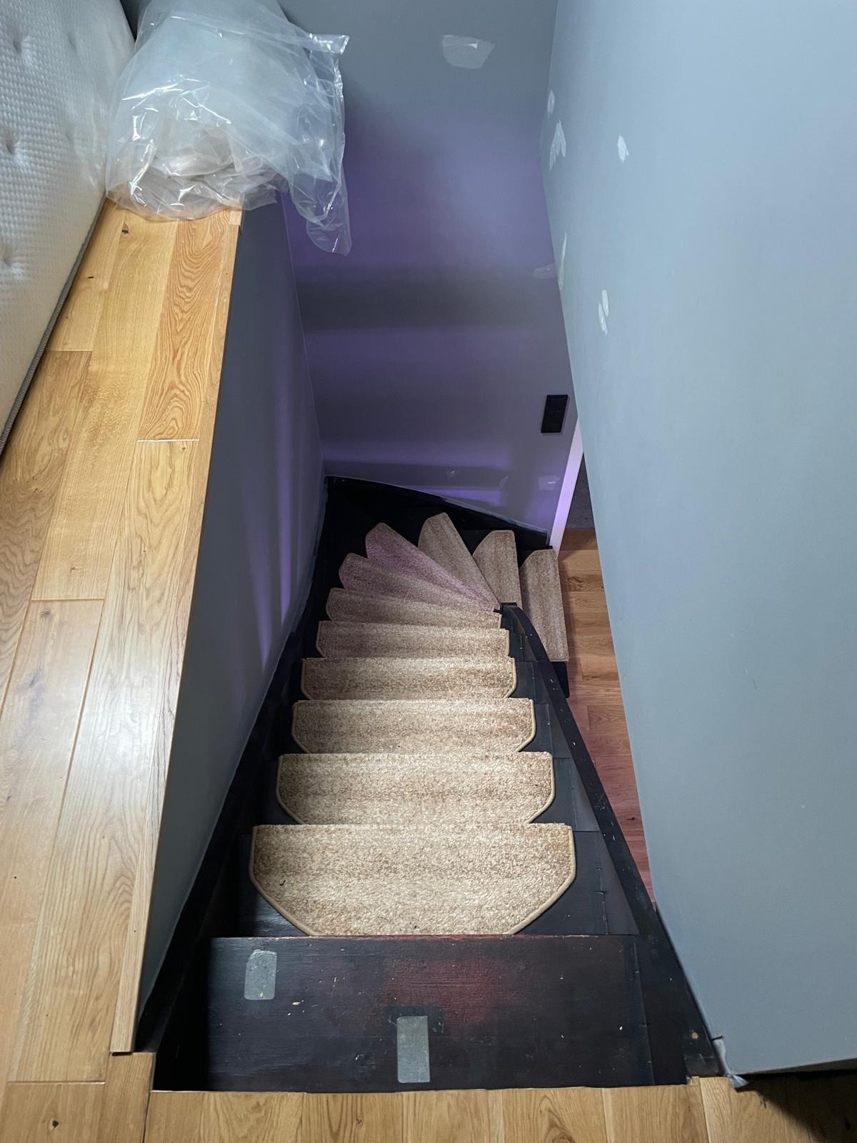 Wysoki kąt widzenia na schody spiralne z wykładziną dywanową w odcieniach beżu, prowadzące w dół. Ściany szare, podłoga drewniana. Widoczne ślady po remoncie.