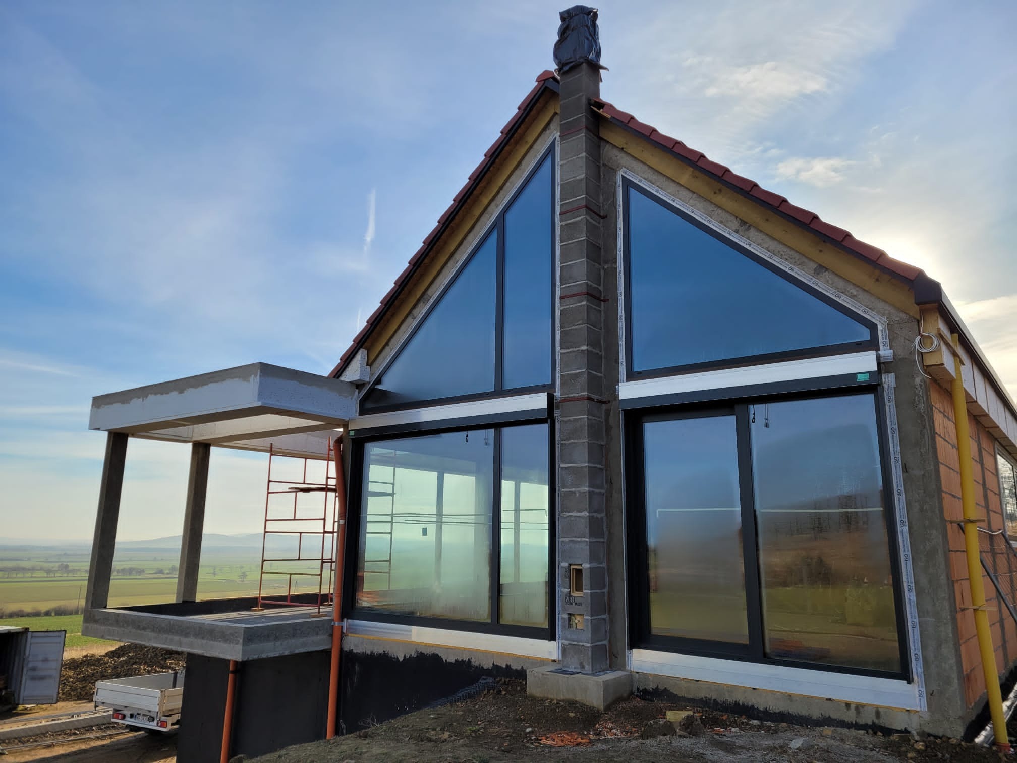 Nowoczesny dom z dużymi oknami aluminiowymi, widok z zewnątrz, w tle krajobraz. Widoczne rusztowanie i elementy konstrukcyjne w trakcie budowy.