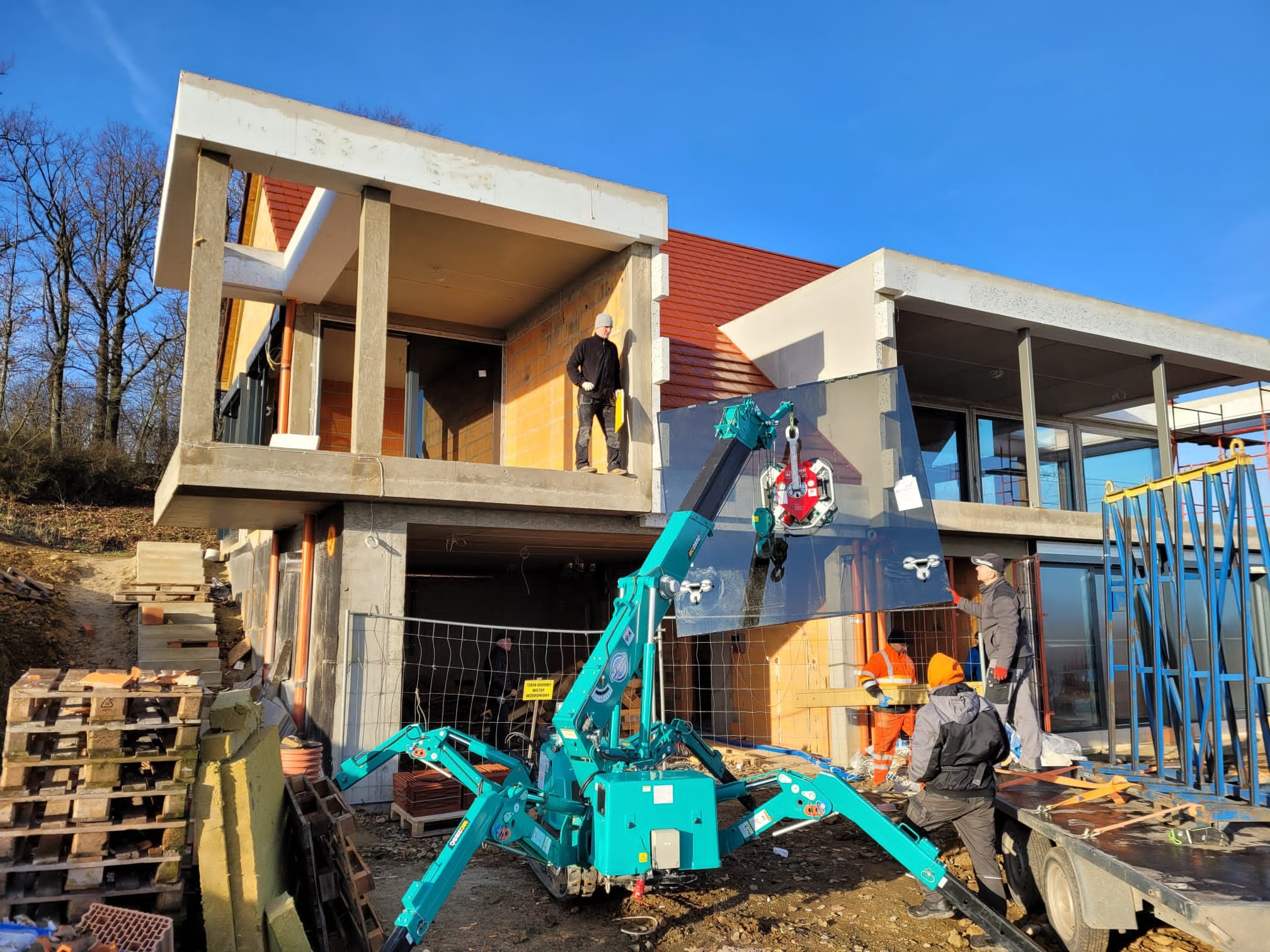 Montaż dużej szyby okiennej za pomocą specjalistycznego dźwigu na budowie domu. Widoczni pracownicy w strojach roboczych. Słoneczny dzień.