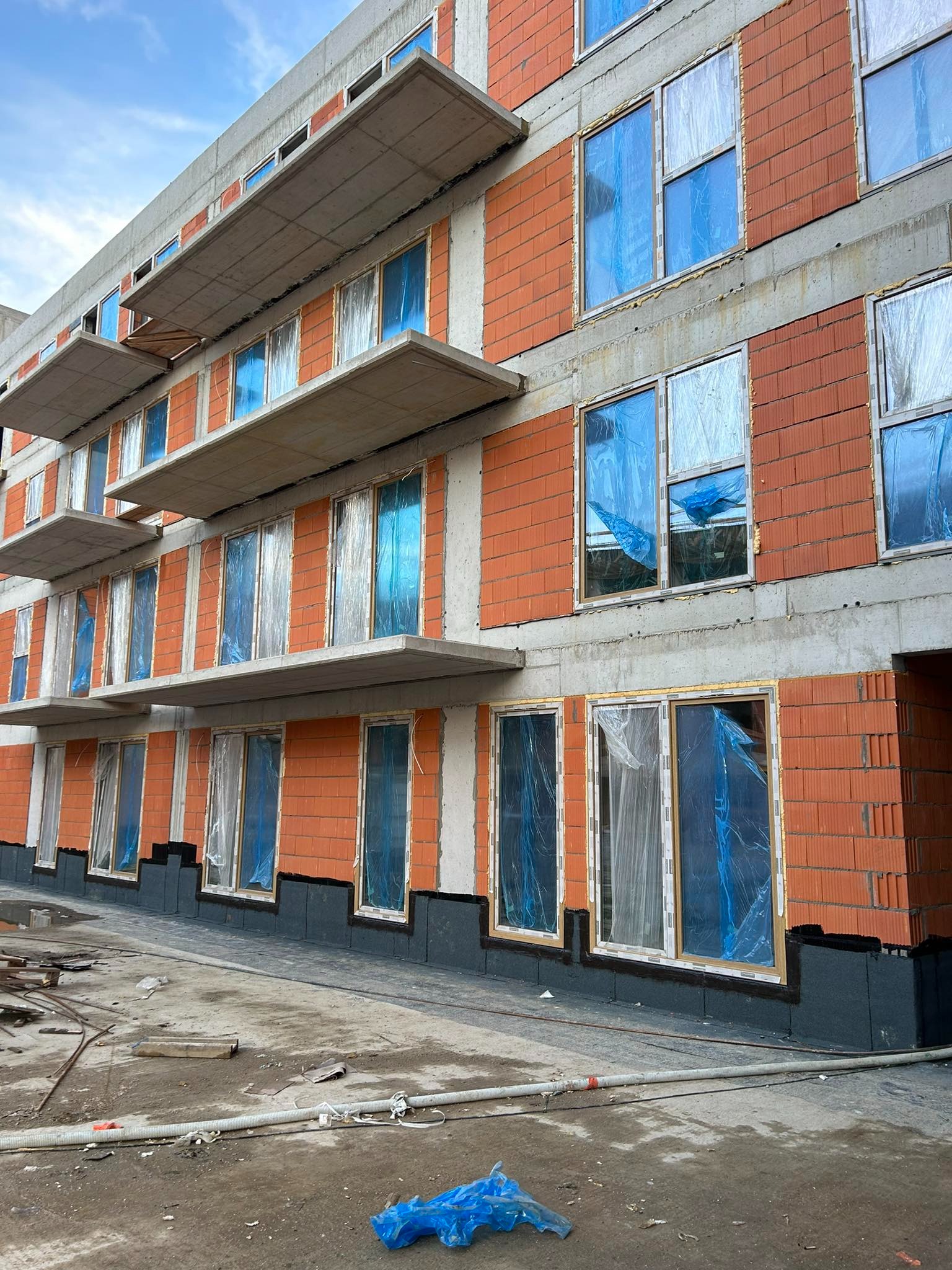 Budynek w trakcie montażu okien, zabezpieczonych folią. Widoczne elementy konstrukcyjne i materiały budowlane. Ujęcie z dołu, w tle błękitne niebo.