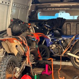NAOMI TRANSPORT - Dwa motocykle crossowe, pomarańczowy i niebieski, zabezpieczone pasami wewnątrz białego busa. Widoczny kask, kanister i narzędzia.