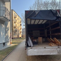 NAOMI TRANSPORT - Otwarta paka samochodu dostawczego z rzeczami do przeprowadzki na tle bloku mieszkalnego i chodnika z kostki. Częściowo widoczne drzewa bez liści.