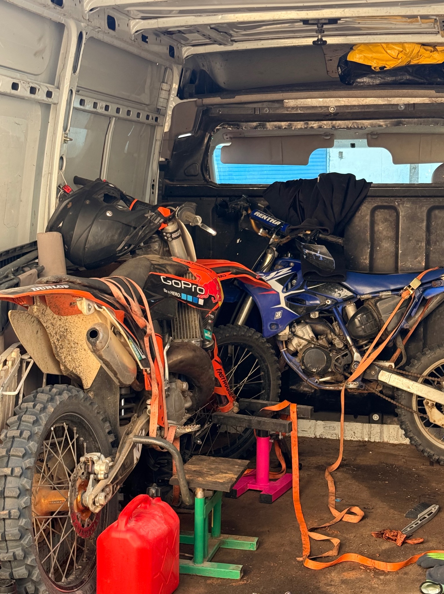 Dwa motocykle crossowe, pomarańczowy i niebieski, zabezpieczone pasami wewnątrz białego busa. Widoczny kask, kanister i narzędzia.
