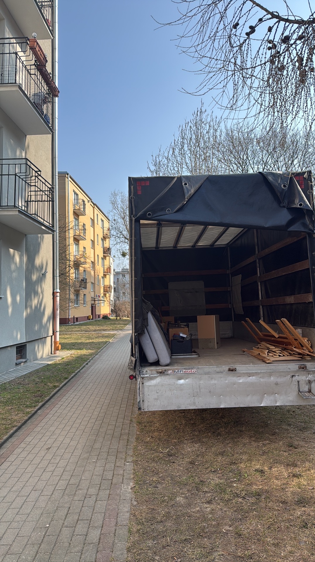 Otwarta paka samochodu dostawczego z rzeczami do przeprowadzki na tle bloku mieszkalnego i chodnika z kostki. Częściowo widoczne drzewa bez liści.