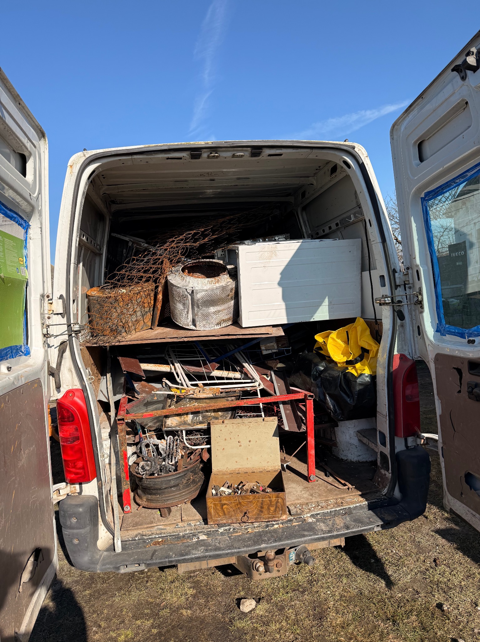 Otwarty furgon wypełniony różnorodnymi przedmiotami przeznaczonymi do utylizacji, w tym metalowymi elementami, sprzętem AGD i workami z odpadami. Widoczne ślady użytkowania pojazdu.