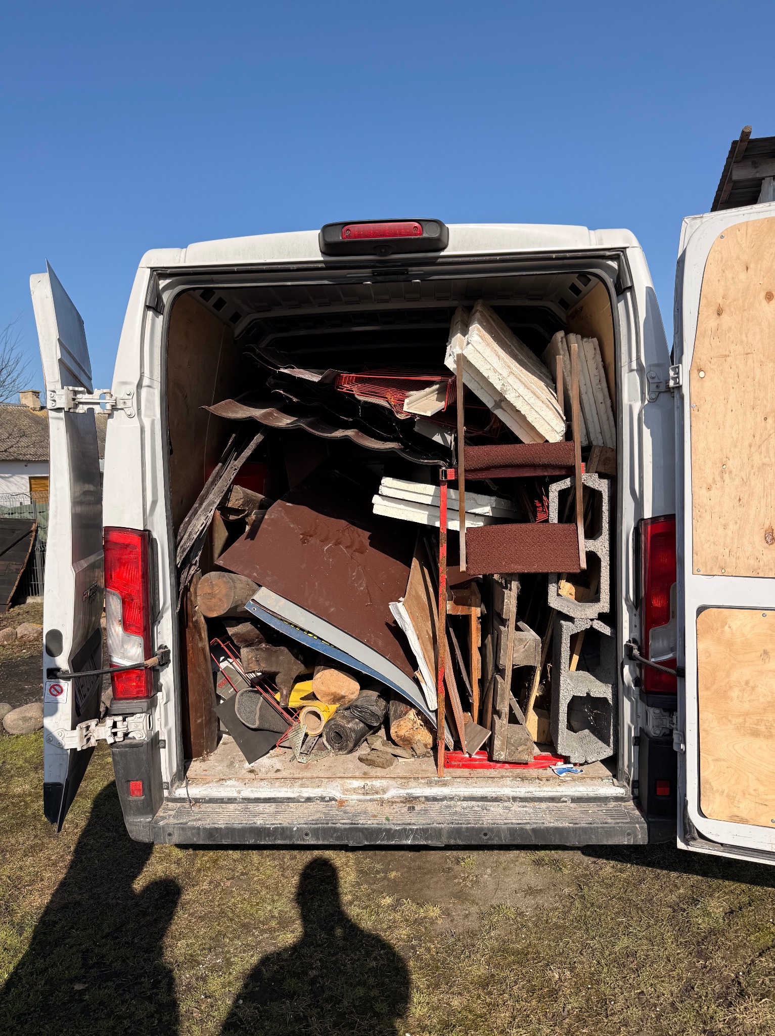 Otwarty bagażnik białego vana wypełniony różnorodnymi przedmiotami, w tym gruzem, drewnem i metalowymi elementami. Widoczny cień osoby na trawie.
