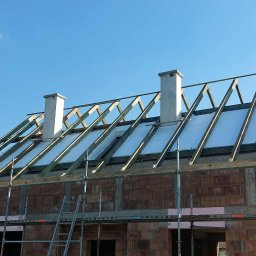 Budowa domu z cegły: widok na konstrukcję dachu z izolacją, kominy i rusztowania na tle błękitnego nieba. Etap prac dekarskich w słoneczny dzień.