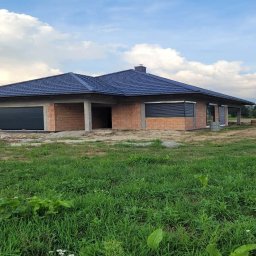 Niedokończony dom jednorodzinny z ciemnoniebieskim dachem z dachówki ceramicznej, widoczny z poziomu trawy w słoneczny dzień. Częściowo otwarte okna.