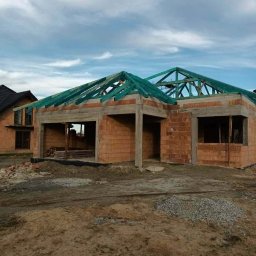 Surowy stan domu jednorodzinnego z cegły, z widoczną konstrukcją dachu z impregnowanego drewna na tle pochmurnego nieba. Etap budowy.