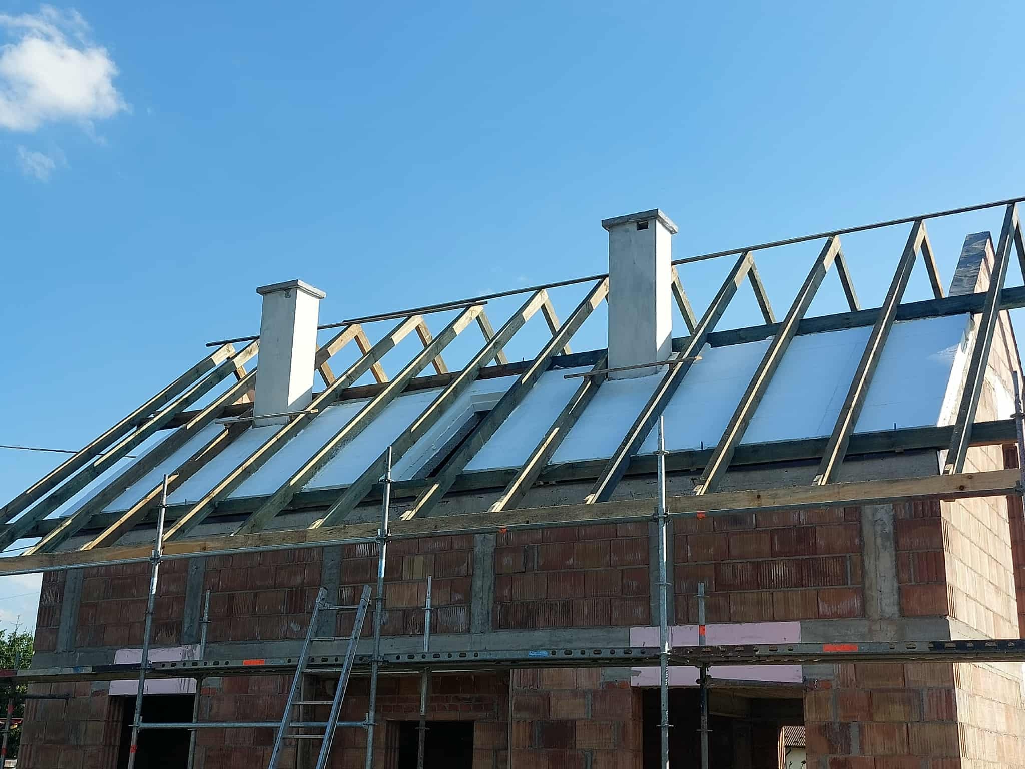 Budowa domu z cegły: widok na konstrukcję dachu z izolacją, kominy i rusztowania na tle błękitnego nieba. Etap prac dekarskich w słoneczny dzień.