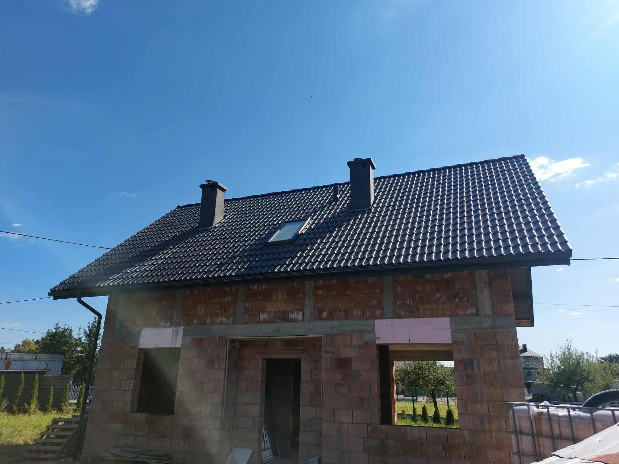 Dom w trakcie budowy z nowym, czarnym dachem z dachówki ceramicznej i zamontowanymi rynnami. Widoczne kominy i okno dachowe. Mur z czerwonej cegły.