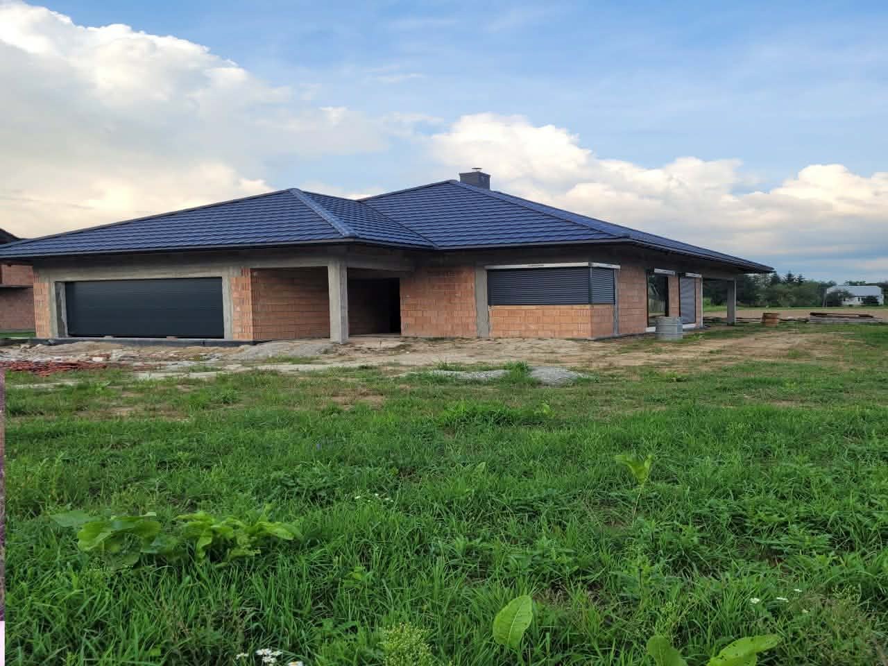 Niedokończony dom jednorodzinny z ciemnoniebieskim dachem z dachówki ceramicznej, widoczny z poziomu trawy w słoneczny dzień. Częściowo otwarte okna.