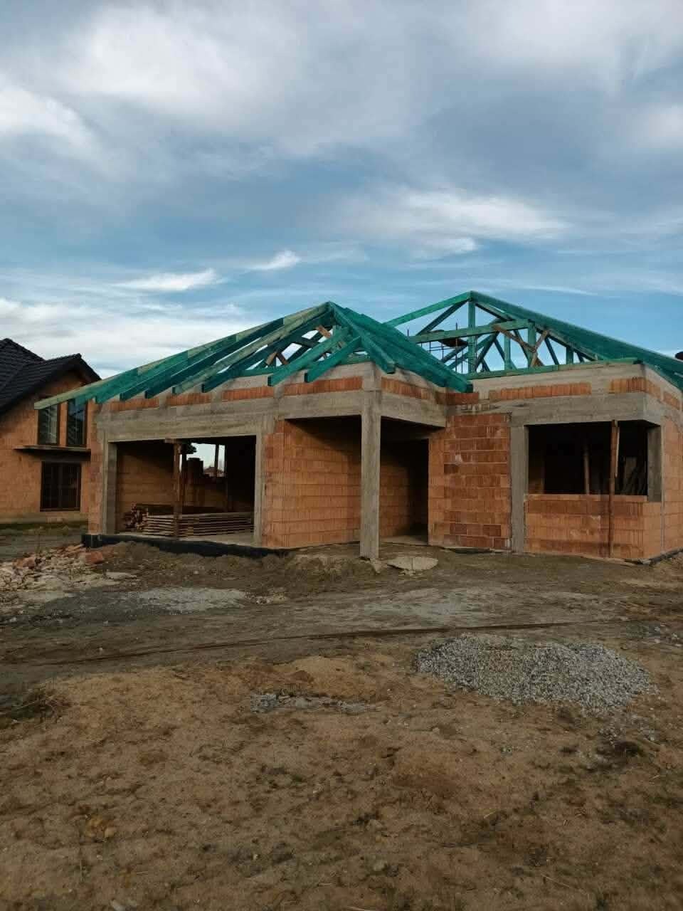 Surowy stan domu jednorodzinnego z cegły, z widoczną konstrukcją dachu z impregnowanego drewna na tle pochmurnego nieba. Etap budowy.