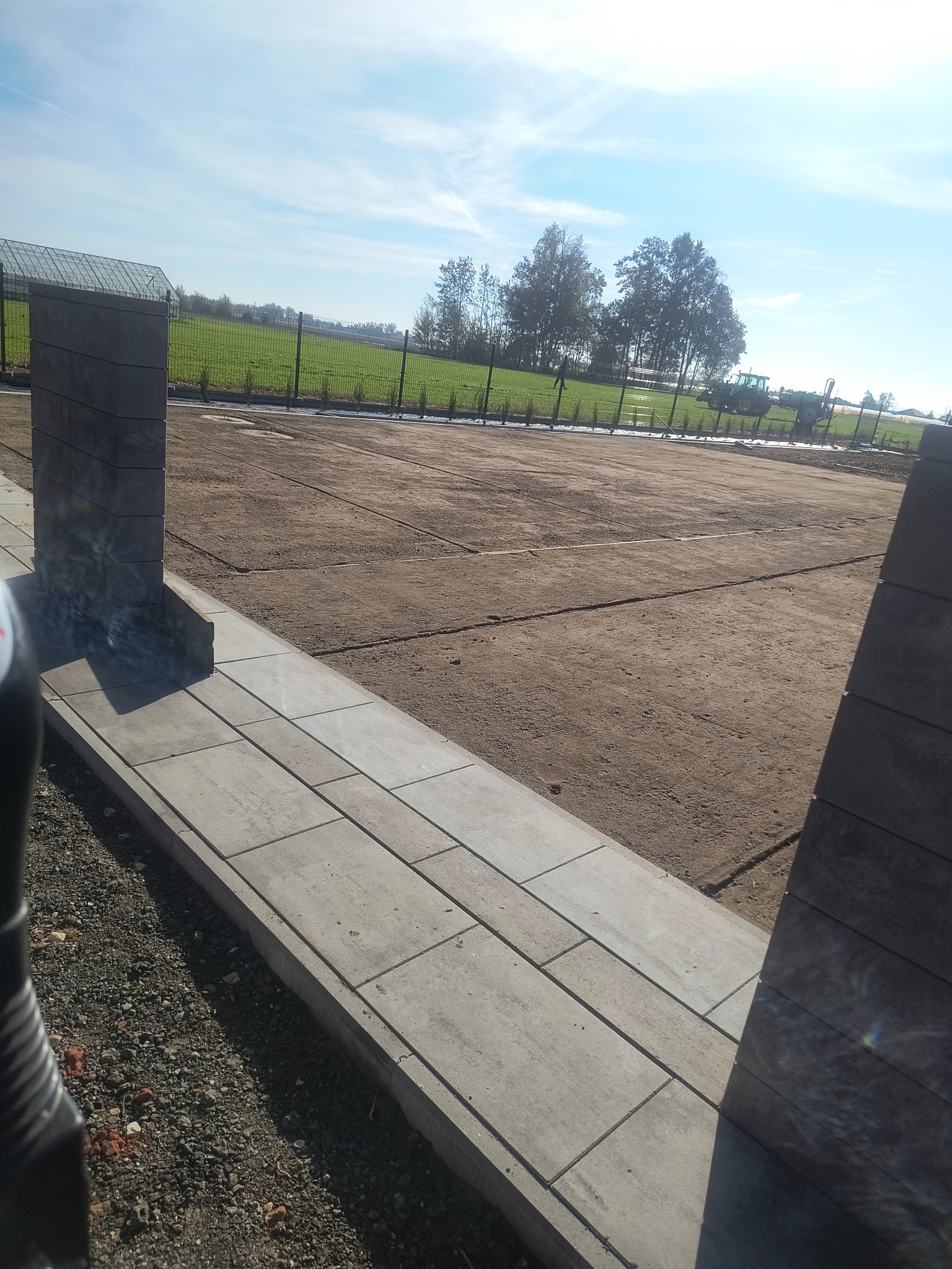 Przygotowany teren pod trawnik z widocznymi śladami po maszynach, obrzeża z kostki i słupki ogrodzeniowe. W tle zielone pole i zabudowania.
