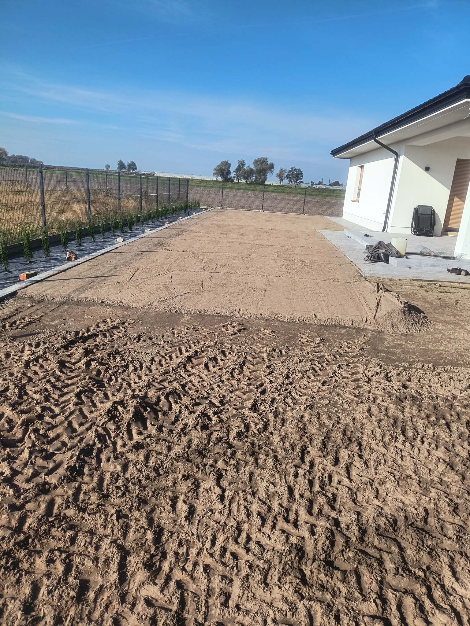 Przygotowany teren pod trawnik przy domu jednorodzinnym. Widoczne ślady opon i wyrównana powierzchnia piasku. W tle ogrodzenie i zieleń.