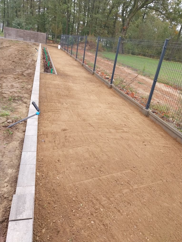 Przygotowany teren pod trawnik z rolki, obrzeża z kostki, widoczny system nawadniania i ogrodzenie panelowe. Perspektywa wzdłuż działki.