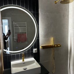 MTM Renowacje - Nowoczesna łazienka w Białymstoku z czarno-białymi płytkami, okrągłym lustrem z podświetleniem LED i złotą armaturą. Minimalistyczny design.