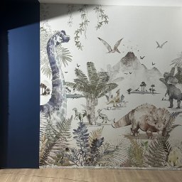 MTM Renowacje - Ściana z tapetą w motywy prehistoryczne: dinozaury, roślinność, wulkan. Fragment niebieskiej ściany po lewej. Widoczna podłoga i listwa przypodłogowa z nazwą producenta tapety.