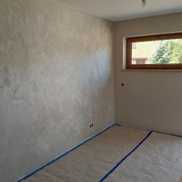 MTM Renowacje - Pokój po malowaniu ścian z efektem dekoracyjnym, zabezpieczona podłoga, okno z widokiem na zieleń, surowa żarówka. Neutralna kolorystyka wnętrza.